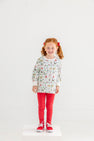 A smiling young girl with red hair in pigtails stands on a white platform, wearing The Beaufort Bonnet Company’s Cassidy Comfy Crewneck - 12 Days of Christmas, paired with red pants, red bows in her hair, and red sneakers, against a plain white background.