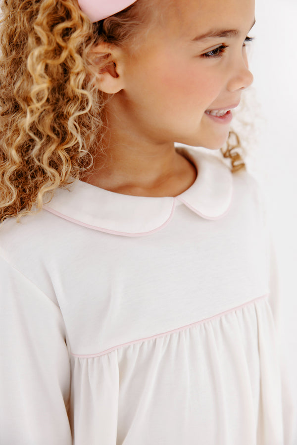 A young girl with curly blonde hair wears The Beaufort Bonnet Company’s Maude’s A Line Top in Palmetto Pearl & Palm Beach Pink, smiling as she looks to the side.