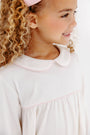 A young girl with curly blonde hair wears The Beaufort Bonnet Company’s Maude’s A Line Top in Palmetto Pearl & Palm Beach Pink, smiling as she looks to the side.