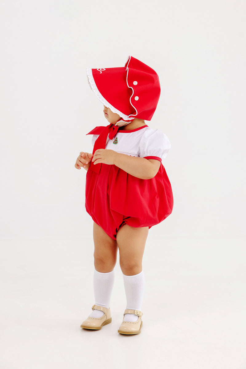A young child stands, facing slightly to the side against a plain white background, wearing The Beaufort Bonnet Company’s Bellefaire Bonnet in Richmond Red with Worth Avenue White Eyelet and Bow, along with a matching red romper featuring white trim, a white shirt, knee-high socks, and beige shoes.
