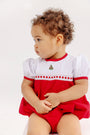 A toddler with curly hair wearing The Beaufort Bonnet Company’s Cindy Lou Bubble in Richmond Red & Worth Avenue White, featuring puffed sleeves and an embroidered Christmas tree, looks to the side against a plain white background.