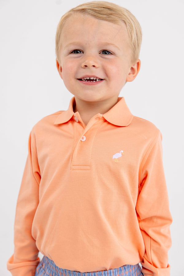 A smiling young child wearing The Beaufort Bonnet Company’s Long Sleeve Prim & Proper Polo & Onesie in Seashore Sherbet with a multicolor stork logo on the chest, standing against a plain white background.