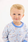 A young child with blonde hair smiles gently, wearing The Beaufort Bonnet Company’s Cassidy Comfy Crewneck – Gourd-geous in Buckhead Blue with Sunrise Blvd Blue accents, adorned with white pumpkins and orange vines—making it the perfect fall piece.