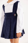 Child wearing a Maude’s Peter Pan Collar Shirt & Onesie (Long Sleeve Woven) - Worth Avenue White under a navy blue corduroy pinafore dress with plaid buttons.