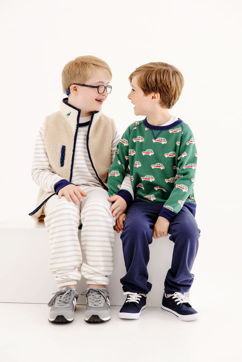 Two young boys sit together, smiling and holding hands. One wears striped pajamas and a fleece vest with a cozy fleece interior, while the other wears The Beaufort Bonnet Company’s Smitty Sweatshirt - Windsor Wagon (Grier Green) with Nantucket Navy, featuring a playful car design. Both look happy and are sitting against a white background.
