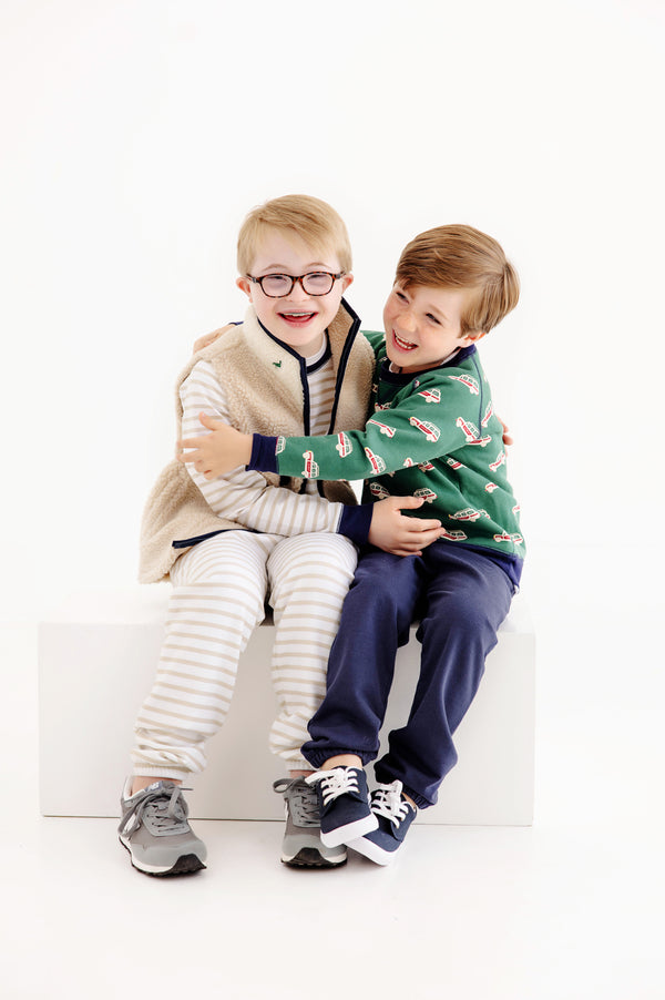 Two young boys sit on a white bench hugging and smiling. One wears glasses, a beige vest, and striped pajamas; the other sports The Beaufort Bonnet Company’s Cassidy & Gates Sweat Set in Keeneland Khaki Stripe & Nantucket Navy. Both look happy and cheerful against a plain background, perfectly displaying comfortable fall loungewear.