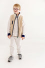 A smiling young boy with short blonde hair and glasses stands against a plain white background, wearing cozy fall loungewear from The Beaufort Bonnet Company. He is dressed in the Cassidy & Gates Sweat Set in Keeneland Khaki Stripe & Nantucket Navy, paired with gray sneakers.