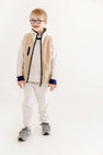 A smiling young boy with short blonde hair and glasses stands against a plain white background, wearing cozy fall loungewear from The Beaufort Bonnet Company. He is dressed in the Cassidy & Gates Sweat Set in Keeneland Khaki Stripe & Nantucket Navy, paired with gray sneakers.
