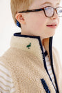 A child with light hair and glasses smiles while wearing The Beaufort Bonnet Company’s Van Camp Vest in Keeneland Khaki Sherpa with Grier Green, featuring a bird patch on the collar and layered over a striped shirt—a perfect fall layering piece.