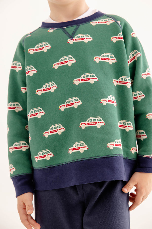 A child wears The Beaufort Bonnet Company’s Smitty Sweatshirt in Windsor Wagon (Grier Green) with Nantucket Navy, featuring a repeating red and white car print, navy cuffs, and a cozy fleece interior, paired with navy pants. The child’s face is not visible.