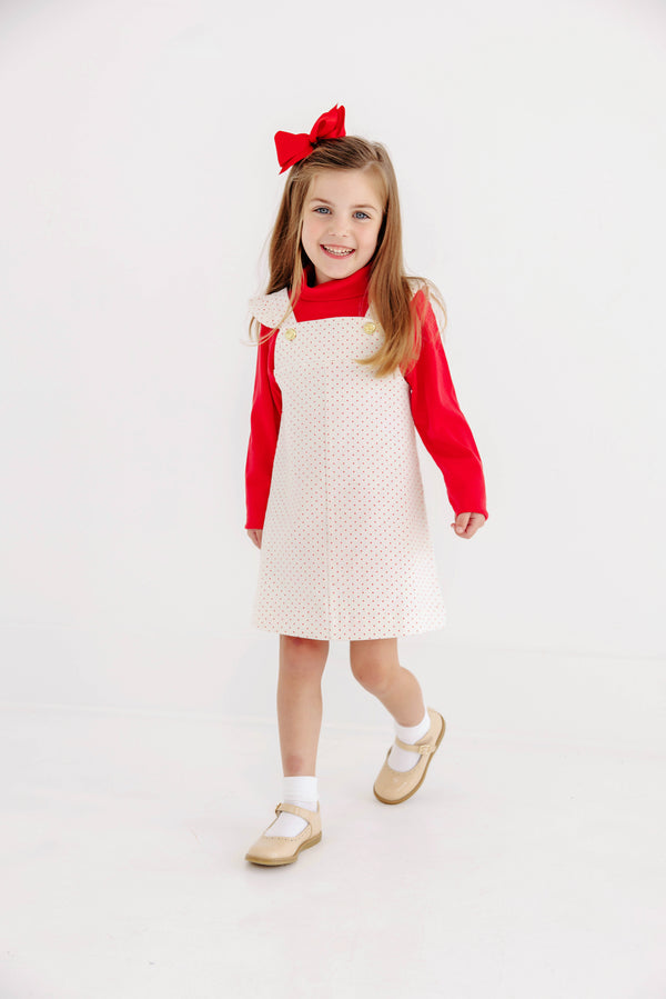 A young girl with long hair wears The Beaufort Bonnet Company’s Mitzy Sue Slacks in Richmond Red, paired with a white pinafore dress featuring yellow buttons, white socks, tan shoes, and a large red bow; she smiles as she walks toward the camera against a plain white background.