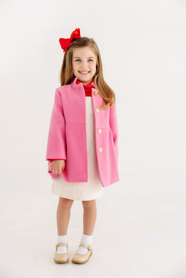 A young girl smiles while wearing The Beaufort Bonnet Company’s Penelope Peacoat in Hamptons Hot Pink with Richmond Red, paired with a white dress featuring small dots, white socks, tan shoes, and a red bow in her hair. The background is plain and white.