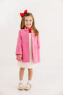 A young girl smiles while wearing The Beaufort Bonnet Company’s Penelope Peacoat in Hamptons Hot Pink with Richmond Red, paired with a white dress featuring small dots, white socks, tan shoes, and a red bow in her hair. The background is plain and white.