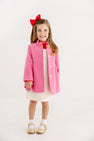 A young girl smiles while wearing The Beaufort Bonnet Company’s Penelope Peacoat in Hamptons Hot Pink with Richmond Red, paired with a white dress featuring small dots, white socks, tan shoes, and a red bow in her hair. The background is plain and white.
