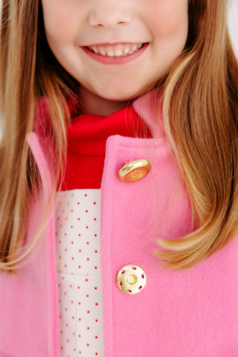 A smiling child wears The Beaufort Bonnet Company’s Penelope Peacoat in Hamptons Hot Pink with Richmond Red, paired with a red scarf and a white shirt with red polka dots; only the lower half of the face and upper torso are visible.