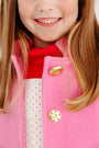 A smiling child wears The Beaufort Bonnet Company’s Penelope Peacoat in Hamptons Hot Pink with Richmond Red, paired with a red scarf and a white shirt with red polka dots; only the lower half of the face and upper torso are visible.