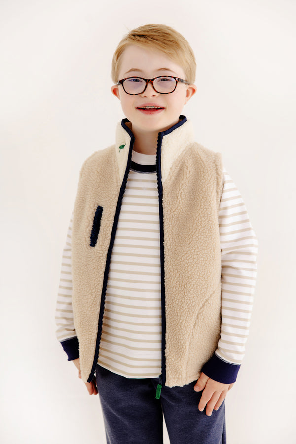 A smiling child with Down syndrome, wearing glasses, a beige fleece vest over a white and gray striped shirt, and The Beaufort Bonnet Company’s Prepletic™ T. Jones Jogger in Nantucket Navy Heathered with Keeneland Khaki Stork, stands against a plain white background.