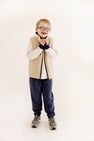 A young boy with glasses smiles joyfully, wearing a striped shirt, beige fleece vest, and gray sneakers, styled in The Beaufort Bonnet Company’s Prepletic™ T. Jones Jogger in Nantucket Navy Heathered with Keeneland Khaki Stork, standing against a plain white background.