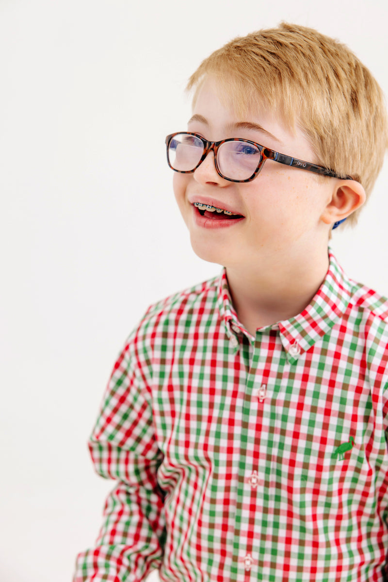 A smiling young boy with glasses and braces is wearing The Beaufort Bonnet Company’s Dean’s List Dress Shirt in Richmond Red & Kiawah Kelly Green Gingham, standing against a plain white background.