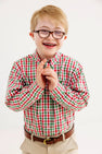A smiling boy with glasses and braces, wearing The Beaufort Bonnet Company’s Dean’s List Dress Shirt in Richmond Red & Kiawah Kelly Green Gingham and khaki pants, stands against a plain white background.