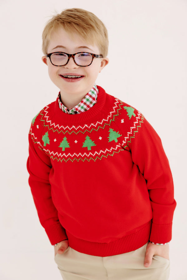 A smiling child with glasses and braces stands against a plain white background, wearing The Beaufort Bonnet Company’s Isaac’s Fair Isle Sweater in Richmond Red with Christmas Trees over a plaid shirt.