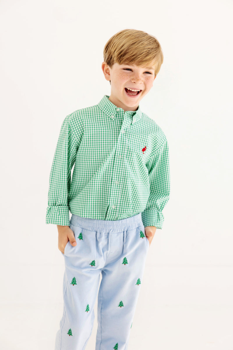 A smiling young boy wears The Beaufort Bonnet Company’s Dean’s List Dress Shirt in Gibson Island Green Mini Gingham with a Richmond Red Stork, paired with light blue pants featuring small green tree prints; his hands are in his pockets as he stands against a plain white background.