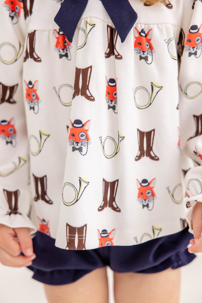 A child wears The Beaufort Bonnet Company’s Long Sleeve Banks Bow Bloomer Set in the Happy Hunting with Palmetto Pearl & Nantucket Navy print. This fall ensemble—featuring a top with whimsical prints of fox faces, brown boots, and yellow French horns paired with navy blue bloomers—is perfect as baby girl autumn clothing. Only the lower torso and hands are visible.