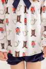 A child wears The Beaufort Bonnet Company’s Long Sleeve Banks Bow Bloomer Set in the Happy Hunting with Palmetto Pearl & Nantucket Navy print. This fall ensemble—featuring a top with whimsical prints of fox faces, brown boots, and yellow French horns paired with navy blue bloomers—is perfect as baby girl autumn clothing. Only the lower torso and hands are visible.
