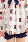 A child wears The Beaufort Bonnet Company’s Long Sleeve Banks Bow Bloomer Set in the Happy Hunting with Palmetto Pearl & Nantucket Navy print. This fall ensemble—featuring a top with whimsical prints of fox faces, brown boots, and yellow French horns paired with navy blue bloomers—is perfect as baby girl autumn clothing. Only the lower torso and hands are visible.