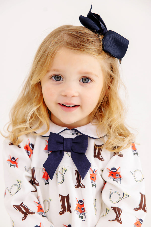 A young child with blonde hair is wearing a navy blue bow in her hair and a matching bow on her shirt, which features a playful Happy Hunting print with boots, foxes, and horns on a white background. She is smiling and facing forward, dressed in the charming Long Sleeve Banks Bow Bloomer Set in Palmetto Pearl & Nantucket Navy from The Beaufort Bonnet Company—a delightful autumn outfit perfect for baby girls this fall.