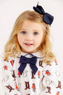 A young child with blonde hair is wearing a navy blue bow in her hair and a matching bow on her shirt, which features a playful Happy Hunting print with boots, foxes, and horns on a white background. She is smiling and facing forward, dressed in the charming Long Sleeve Banks Bow Bloomer Set in Palmetto Pearl & Nantucket Navy from The Beaufort Bonnet Company—a delightful autumn outfit perfect for baby girls this fall.