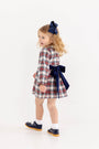 A young girl wearing a Long Sleeve Cindy Lou Sash Dress - Paris Pike Plaid with Nantucket Navy, featuring a large navy blue bow at the waist, a matching hair bow, white socks, and navy blue shoes.