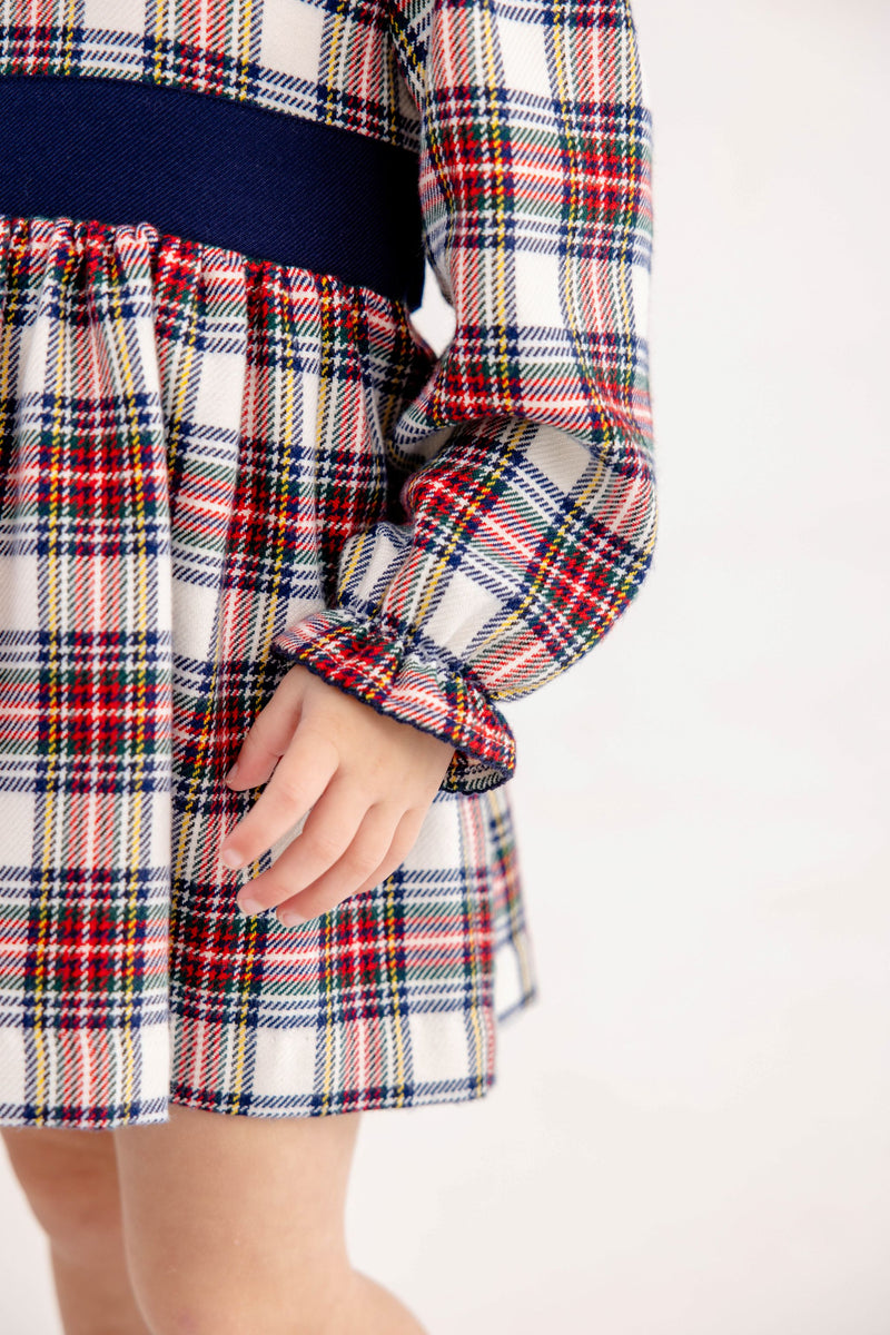 A child wears a Long Sleeve Cindy Lou Sash Dress - Paris Pike Plaid with Nantucket Navy, featuring a navy blue sash at the waist and ruffled sleeve cuffs, perfect as a holiday party dress.