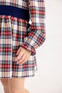 A child wears a Long Sleeve Cindy Lou Sash Dress - Paris Pike Plaid with Nantucket Navy, featuring a navy blue sash at the waist and ruffled sleeve cuffs, perfect as a holiday party dress.