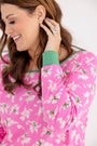 A woman smiles while wearing The Beaufort Bonnet Company’s Long Sleeve Stay in Bed Set in the Bows of Holly with Kiawah Kelly Green colorway, featuring pink floral patterns and green cuffs that evoke a festive pajama style. She is shown from the upper body and in profile, touching her earring. Her brown wavy hair cascades gently, complemented by a ring and earrings.