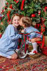 A young girl wearing a Long Sleeve Sandy Smocked Dress - Barrington Blue with Richmond Red Holly kneels beside a toddler on a red tricycle as the child kisses her cheek in front of a decorated Christmas tree.