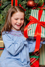 A smiling girl wears a Long Sleeve Sandy Smocked Dress in Barrington Blue with Richmond Red Holly while holding a gift wrapped in green and white striped paper with a large red bow in front of a Christmas tree.