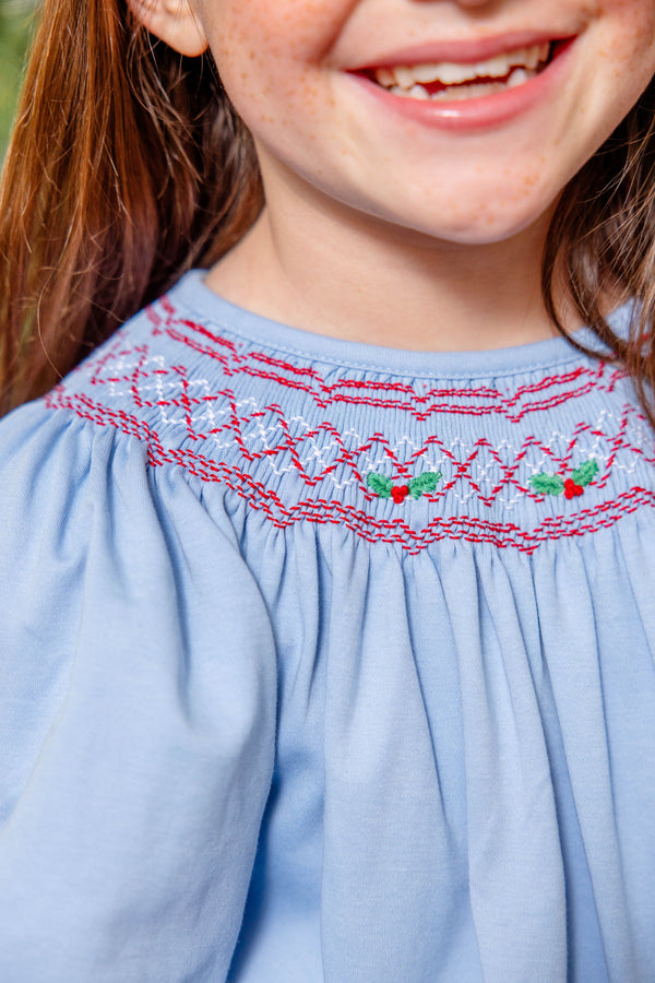 The Long Sleeve Sandy Smocked Dress - Barrington Blue with Richmond Red Holly is a light blue long-sleeve Christmas smocked dress featuring red and white holly berry and leaf patterns across the chest.