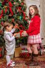 A girl in a red cardigan and a Perrin Skirt - Broadway Play Plaid hands a wrapped gift to a young boy in a grey houndstooth vest and shorts in front of a decorated Christmas tree.