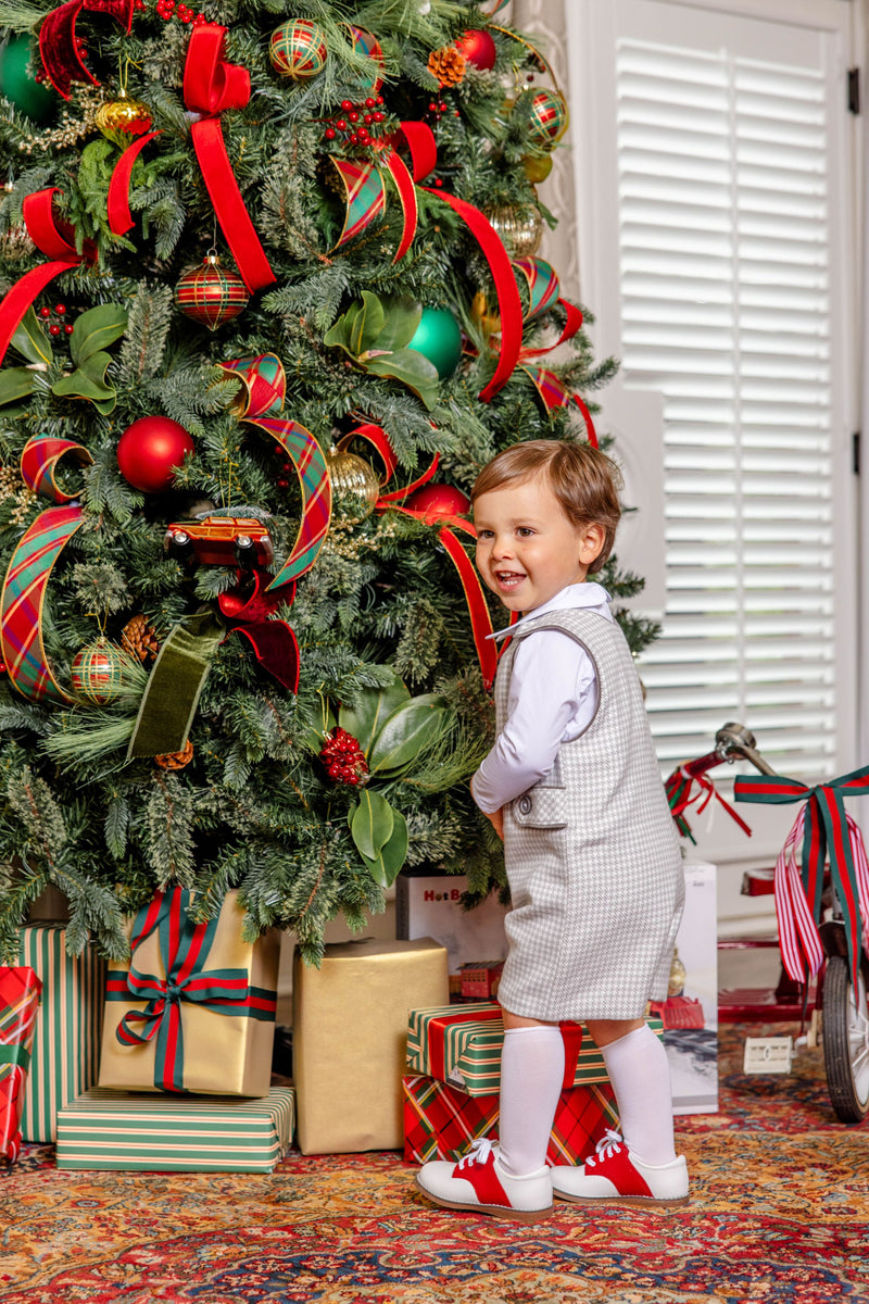 A young boy wearing a Jack Keene Jon Jon - Grantley Gray Houndstooth with Grantley Gray, a white shirt, and red and white shoes stands smiling in front of a decorated Christmas tree for a winter family portrait.