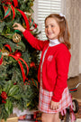 A young girl in a Richmond Red Cambridge Cardigan (Unisex) worn over a white turtleneck and plaid skirt decorates a Christmas tree.