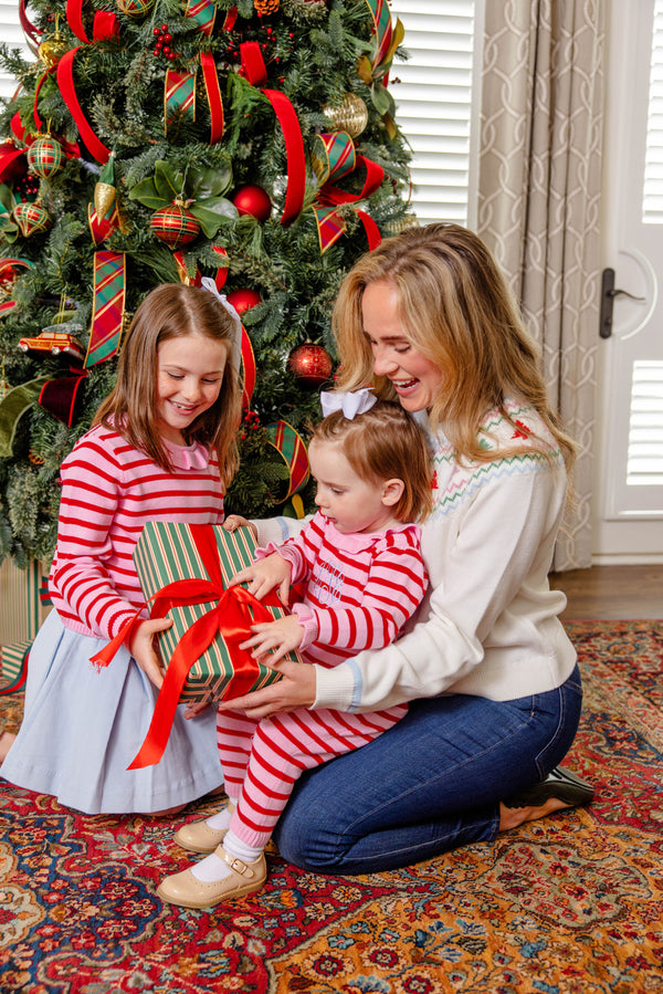 A woman and two young girls, dressed in festive clothes, open a striped gift with a red bow in front of a decorated Christmas tree in a cozy living room. One of the girls is wearing The Beaufort Bonnet Company’s Reece Ruffle Romper in Richmond Red & Pier Party Pink Stripe—the perfect 100% cotton holiday outfit for celebrating together.