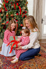 A woman and two young girls, dressed in festive clothes, open a striped gift with a red bow in front of a decorated Christmas tree in a cozy living room. One of the girls is wearing The Beaufort Bonnet Company’s Reece Ruffle Romper in Richmond Red & Pier Party Pink Stripe—the perfect 100% cotton holiday outfit for celebrating together.