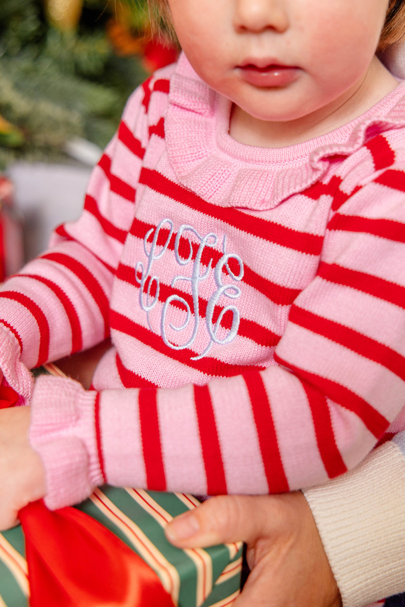 A young child wearing The Beaufort Bonnet Company’s Reece Ruffle Romper in Richmond Red & Pier Party Pink Stripe sits on someone’s lap and holds a wrapped present with green and red stripes, making for the perfect holiday outfit.