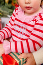 A young child wearing The Beaufort Bonnet Company’s Reece Ruffle Romper in Richmond Red & Pier Party Pink Stripe sits on someone’s lap and holds a wrapped present with green and red stripes, making for the perfect holiday outfit.