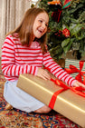 A smiling child wearing The Beaufort Bonnet Company’s Reece Ruffle Sweater in Pier Party Pink & Richmond Red Stripe sits on a carpeted floor by a decorated Christmas tree, holding a large, gold-wrapped present with a red ribbon.