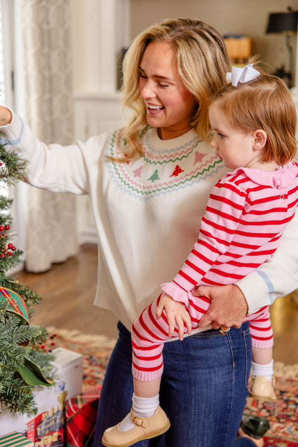 A woman holds a young girl dressed in the Reece Ruffle Romper—Richmond Red & Pier Party Pink Stripe by The Beaufort Bonnet Company—as they decorate a Christmas tree in a cozy living room, with presents arranged beneath the tree.