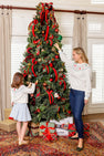 A woman and a child, both wearing The Beaufort Bonnet Company’s Isabelle’s Fair Isle Sweater in Palmetto Pearl with Trees, decorate a Christmas tree adorned with red and green ornaments. Wrapped gifts sit beneath the tree while sunlight pours through the windows, casting a warm glow over their festive holiday wardrobe scene.