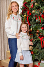 A smiling woman and young girl stand in front of a decorated Christmas tree, both wearing matching white sweaters adorned with a colorful tree and zigzag pattern. The girl, dressed in the Parson Pleated Skirt in Buckhead Blue Corduroy by The Beaufort Bonnet Company, holds a gold ornament as they both look toward the camera.