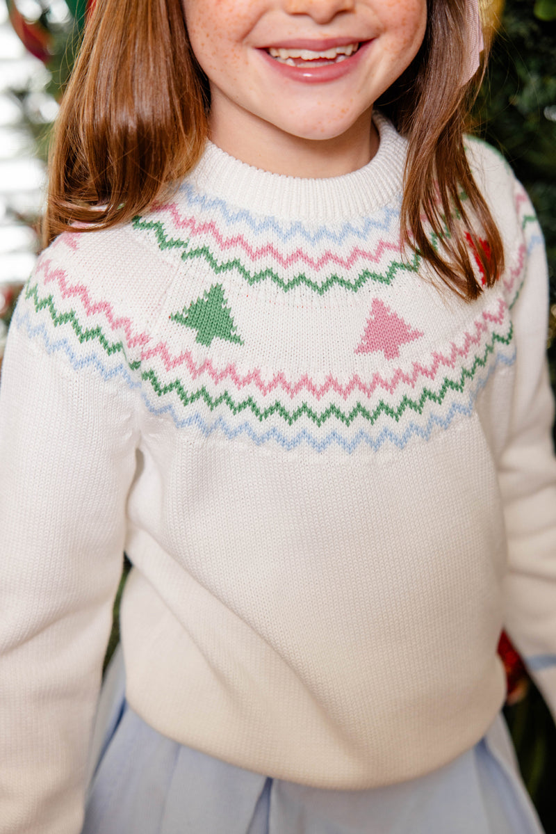 A child smiles in front of a blurred background, dressed in The Beaufort Bonnet Company’s Isabelle’s Fair Isle Sweater—crafted from 100% cotton in the Palmetto Pearl colorway with charming green and pink tree patterns and zigzag designs—paired with a light blue skirt.